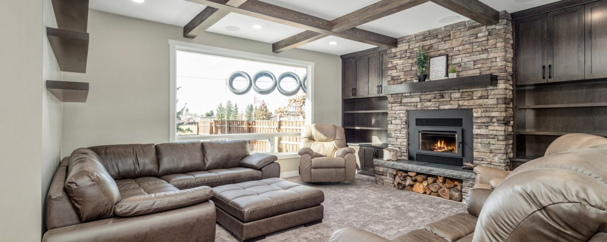 Cozy living room with stone fireplace, exposed wood ceiling beams, built-in cabinetry, leather recliners, and large window with natural light.